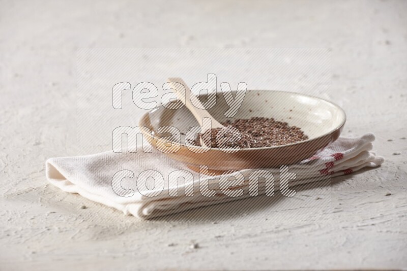 A multicolored pottery plate full of flax seeds with a wooden spoon full of the seeds on a napkin on a textured white flooring