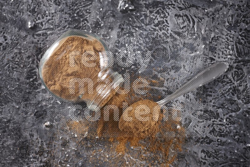 Herbal glass jar full of cinnamon powder flipped and a metal spoon on textured black background