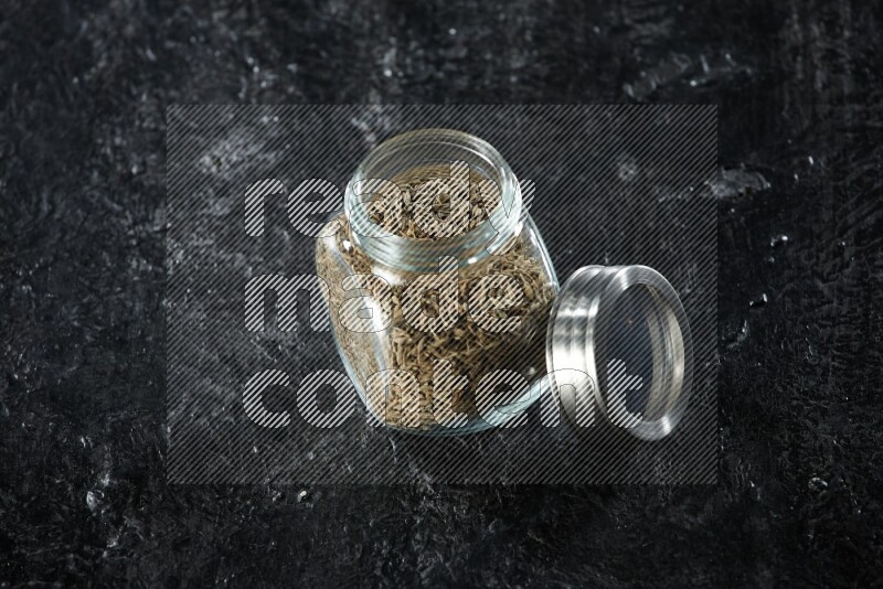 A glass spice jar full of cumin seeds on a textured black flooring