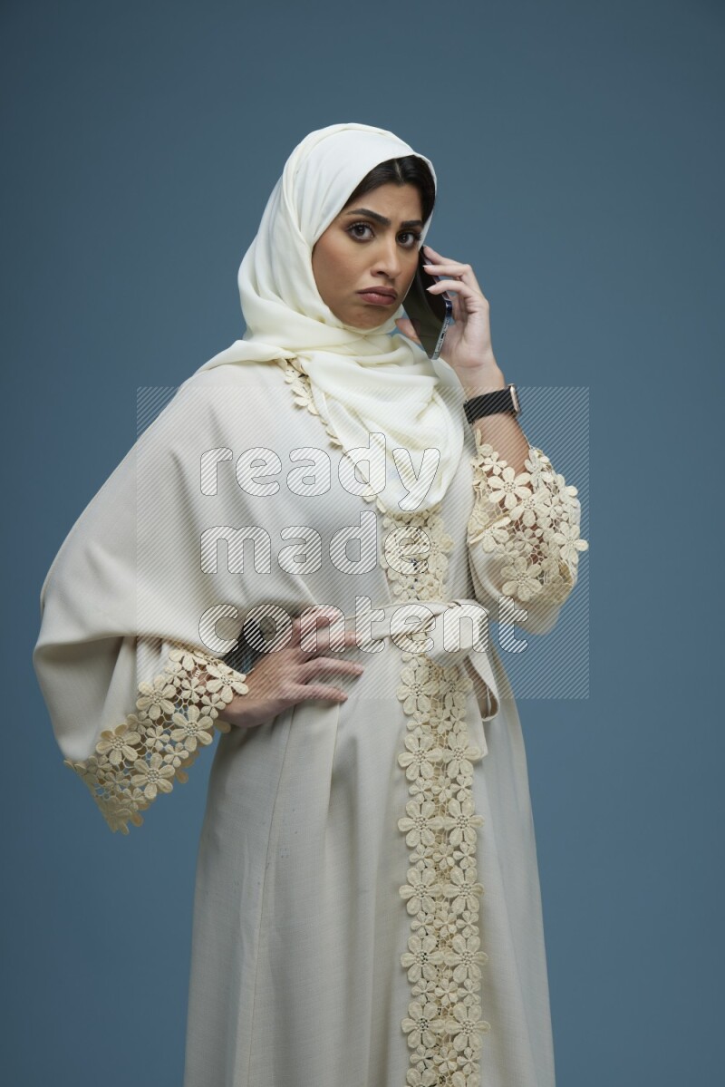 A Saudi woman having a call in a blue background wearing an off-white Abaya Hijab