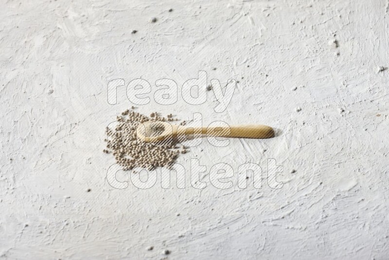 A wooden spoon full of white pepper powder and white pepper beads on textured white flooring