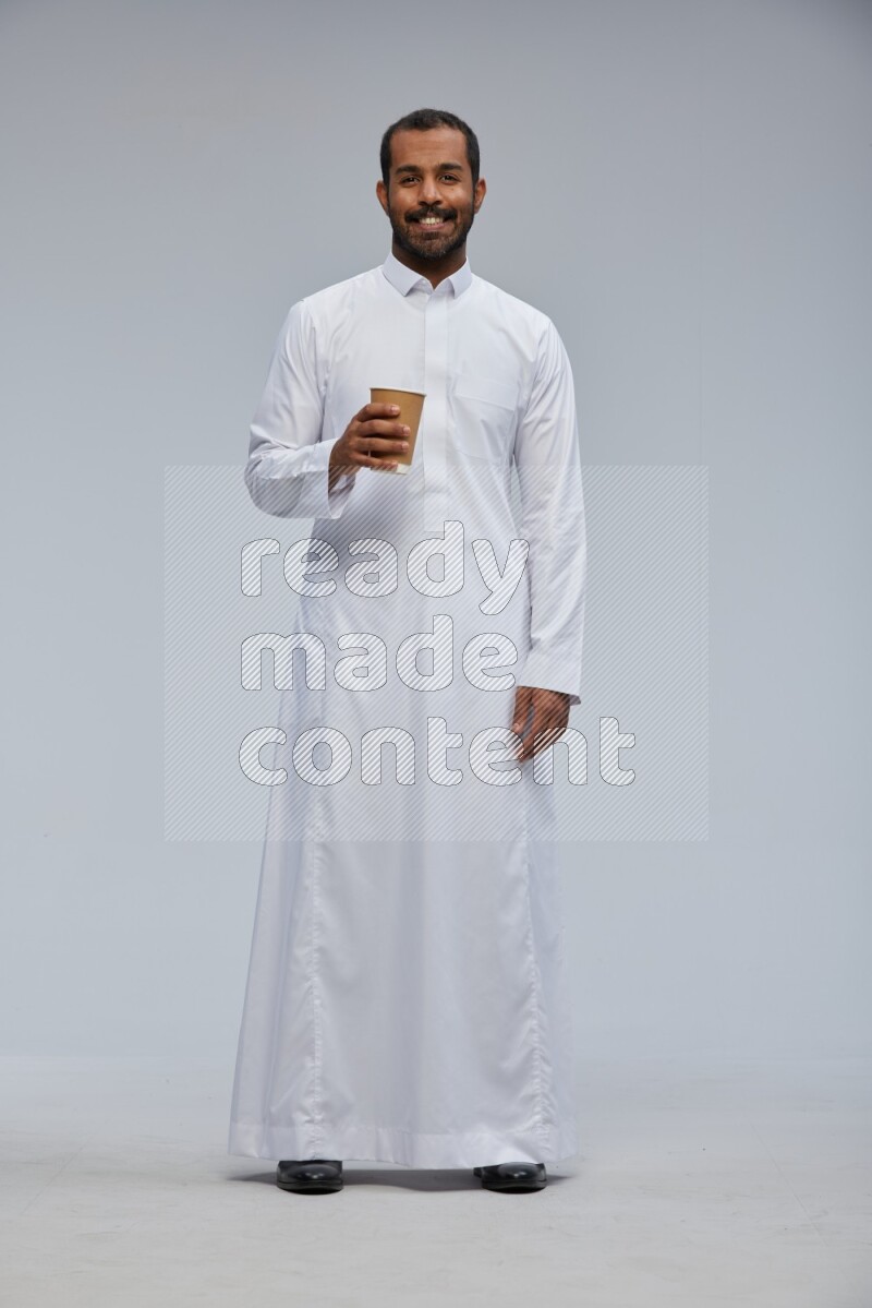 Saudi man wearing thob standing holding paper cup on gray background