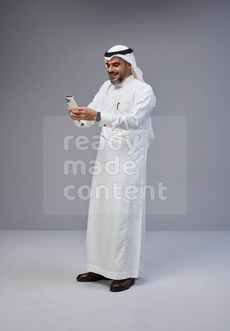 Saudi man Wearing Thob and white Shomag standing texting on phone on Gray background