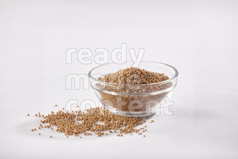 A glass bowl full of mustard seeds and more seeds spread on a white flooring