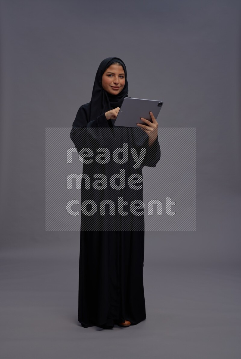 Saudi woman wearing Abaya standing working on tablet on gray background