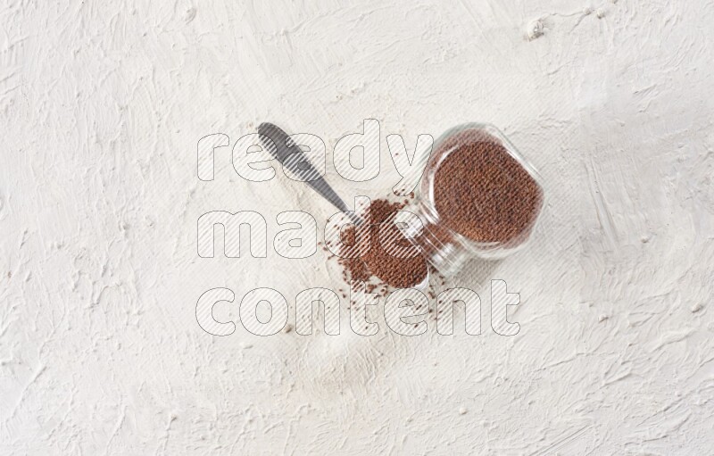 A glass spice jar and a metal spoon full of garden cress seeds and jar is flipped with fallen seeds on a textured white flooring