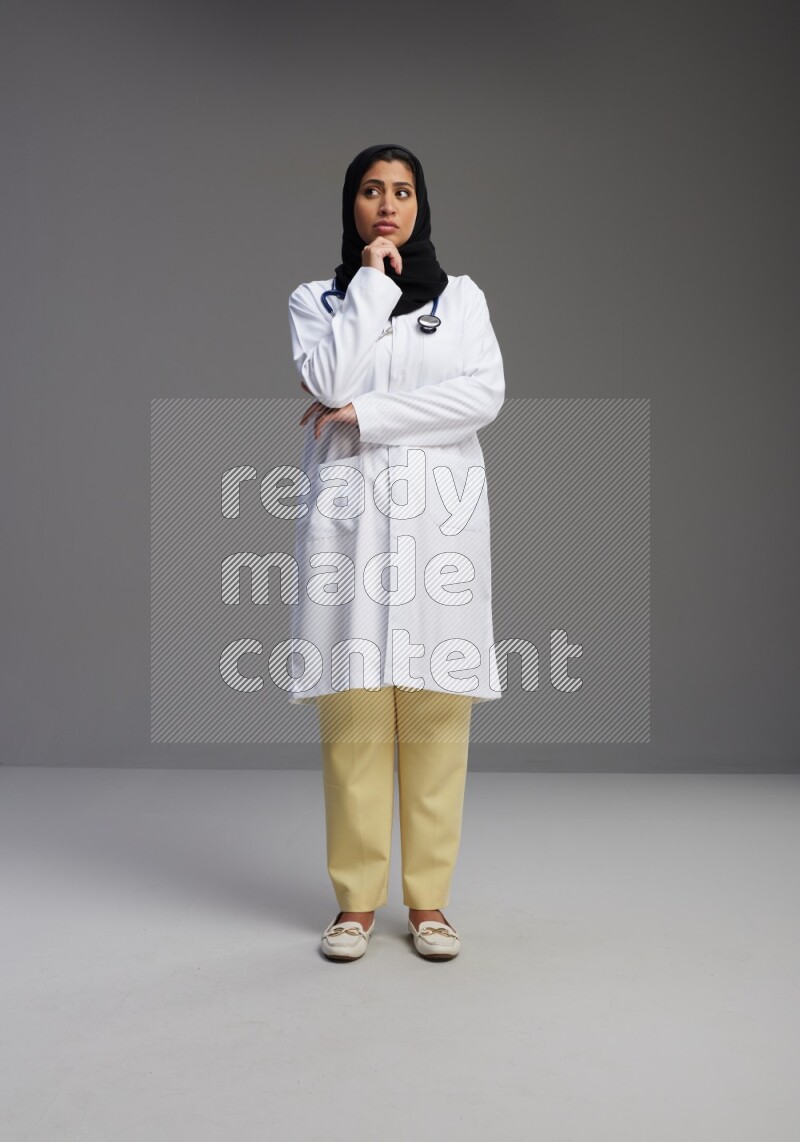 Saudi woman wearing lab coat with stethoscope standing interacting with the camera on Gray background