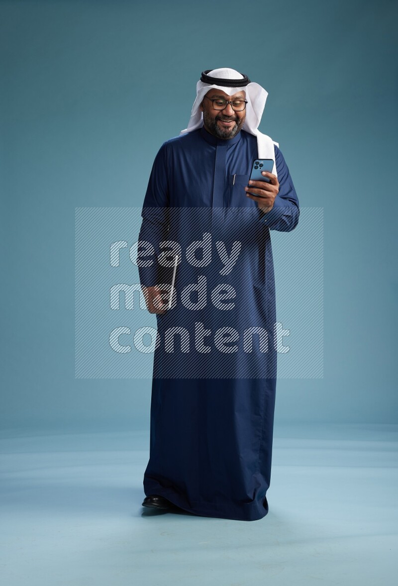 Saudi Man with shimag Standing texting on phone on blue background