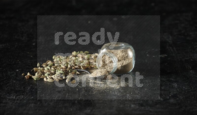 وعاء زجاجي للتوابل مقلوب وملعقة معدنية ممتلئة ببودرة الهيل وحبوب الهيل متناثرة على خلفية سوداء