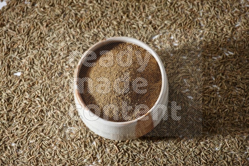 A beige bowl full of cumin powder with cumin seeds beneath it the bowl on a white flooring