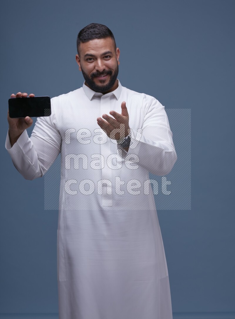 A man Showing his phone in Blue Background wearing Saudi Thob