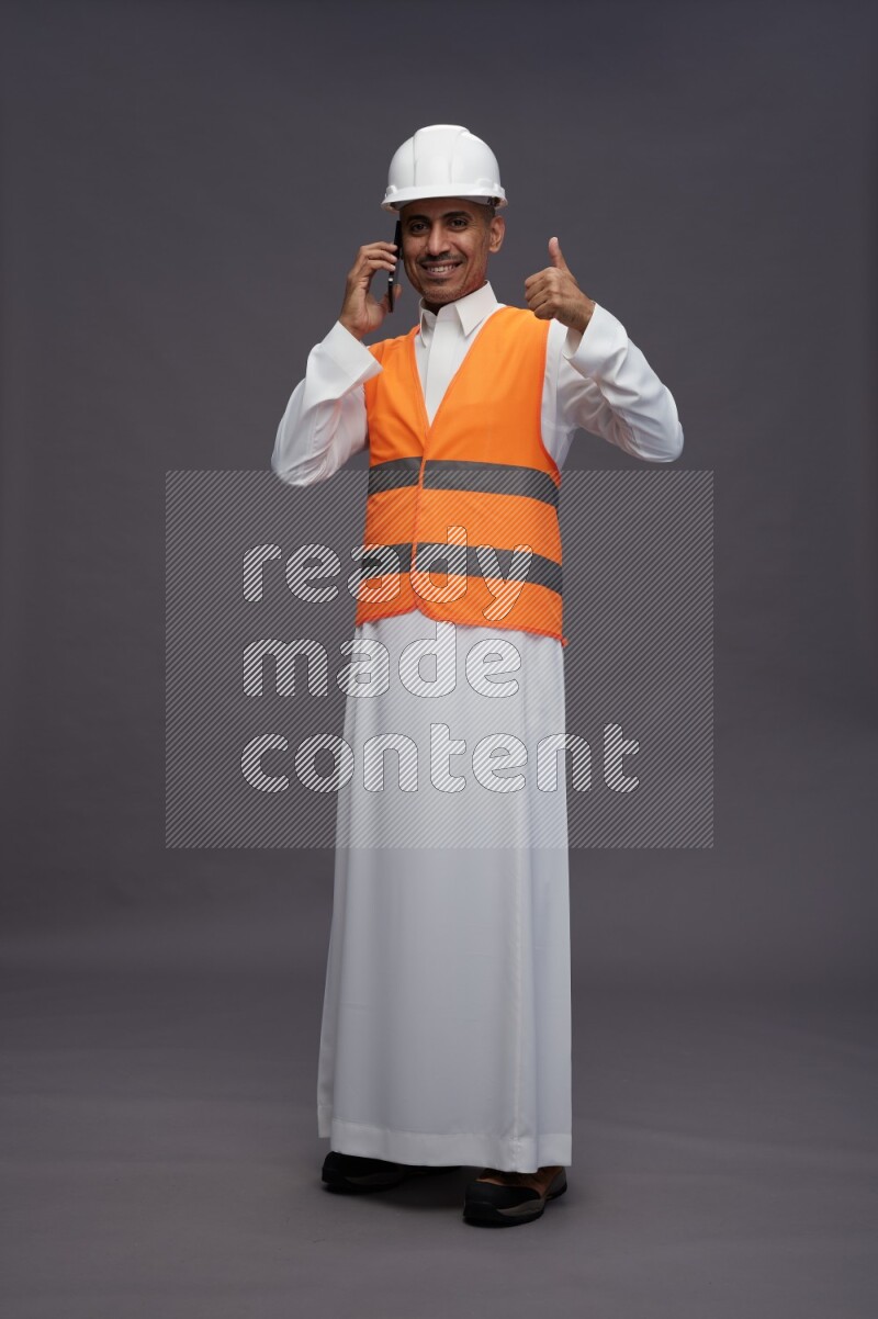 Saudi man wearing thob with engineer vest standing talking on phone on gray background