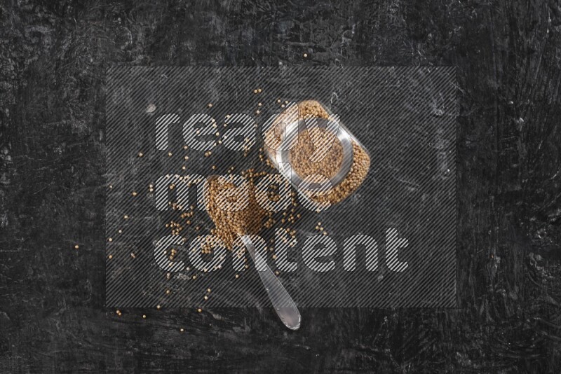 A glass spice jar and a metal spoon full of mustard seeds on a textured black flooring