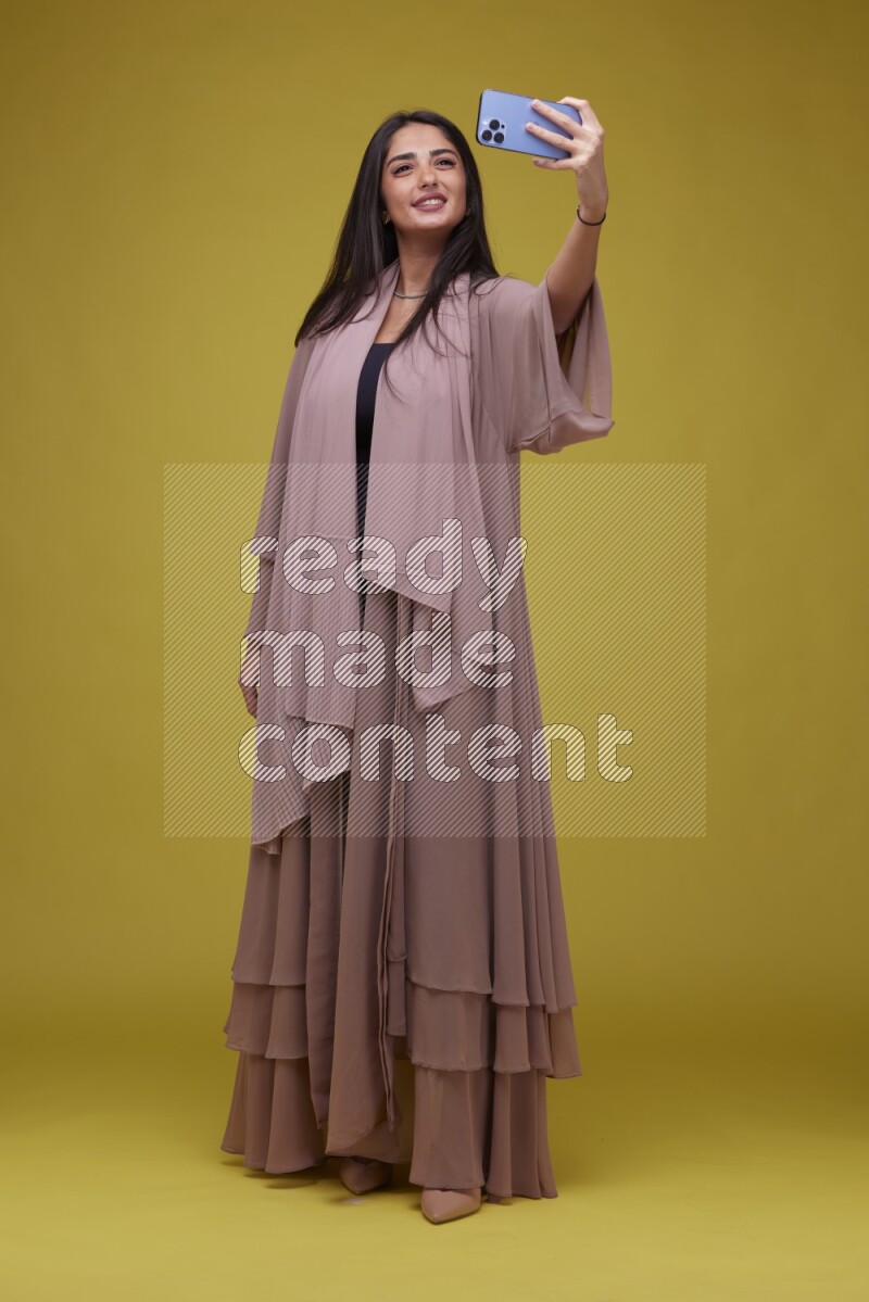 A woman Taking a Selfie on a Yellow Background wearing Brown Abaya