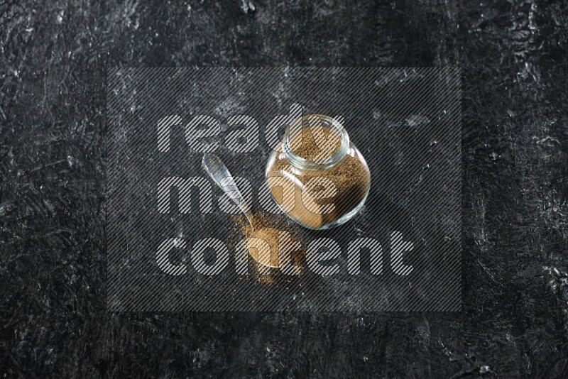 وعاء زجاجي للتوابل وملعقة معدنية ممتلئين ببودرة الكمون على خلفية سوداء