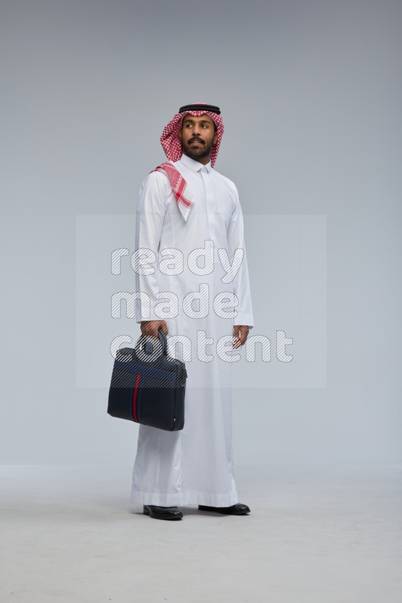 Saudi man Wearing Thob and shomag standing holding bag on Gray background