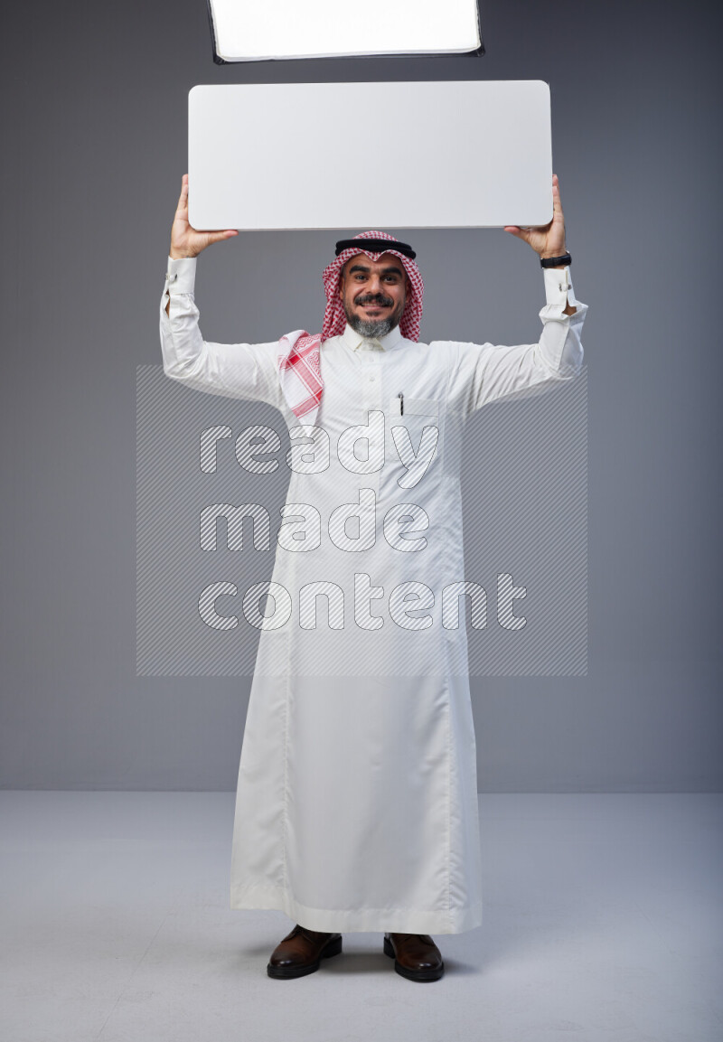 Saudi man Wearing Thob and red Shomag standing holding board on Gray background