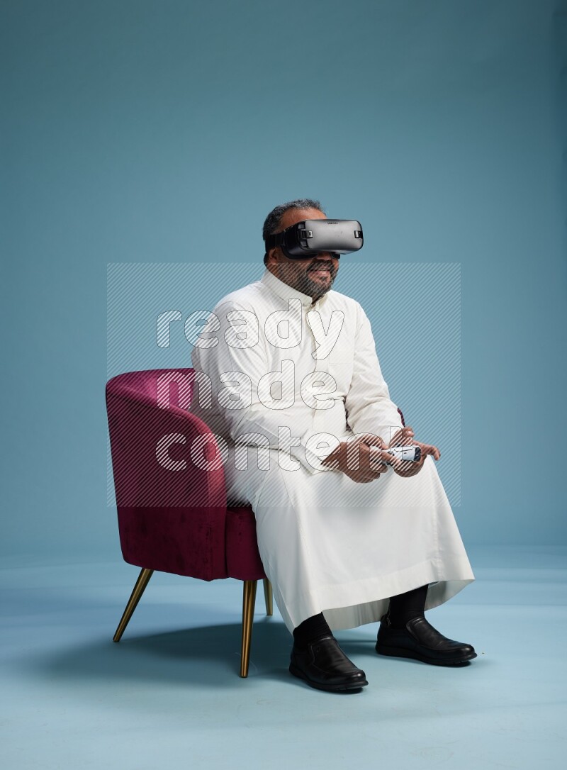 Saudi Man without shimag sitting on chair  gaming with VR and joystick on blue background