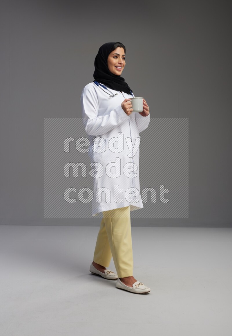 Saudi woman wearing lab coat with stethoscope standing holding mug on Gray background