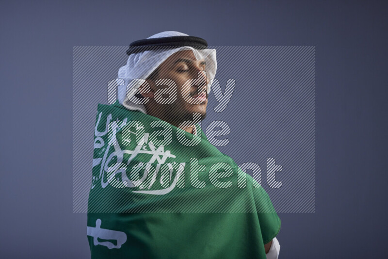 A close-up shot of Saudi man wearing thob and white shomag wrapping big Saudi flag on gray background