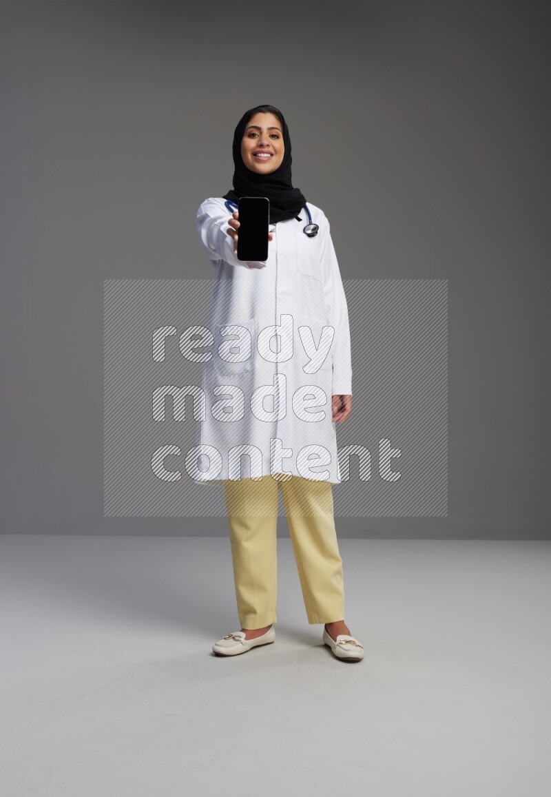 Saudi woman wearing lab coat with stethoscope standing showing phone to camera with sign in the back on Gray background