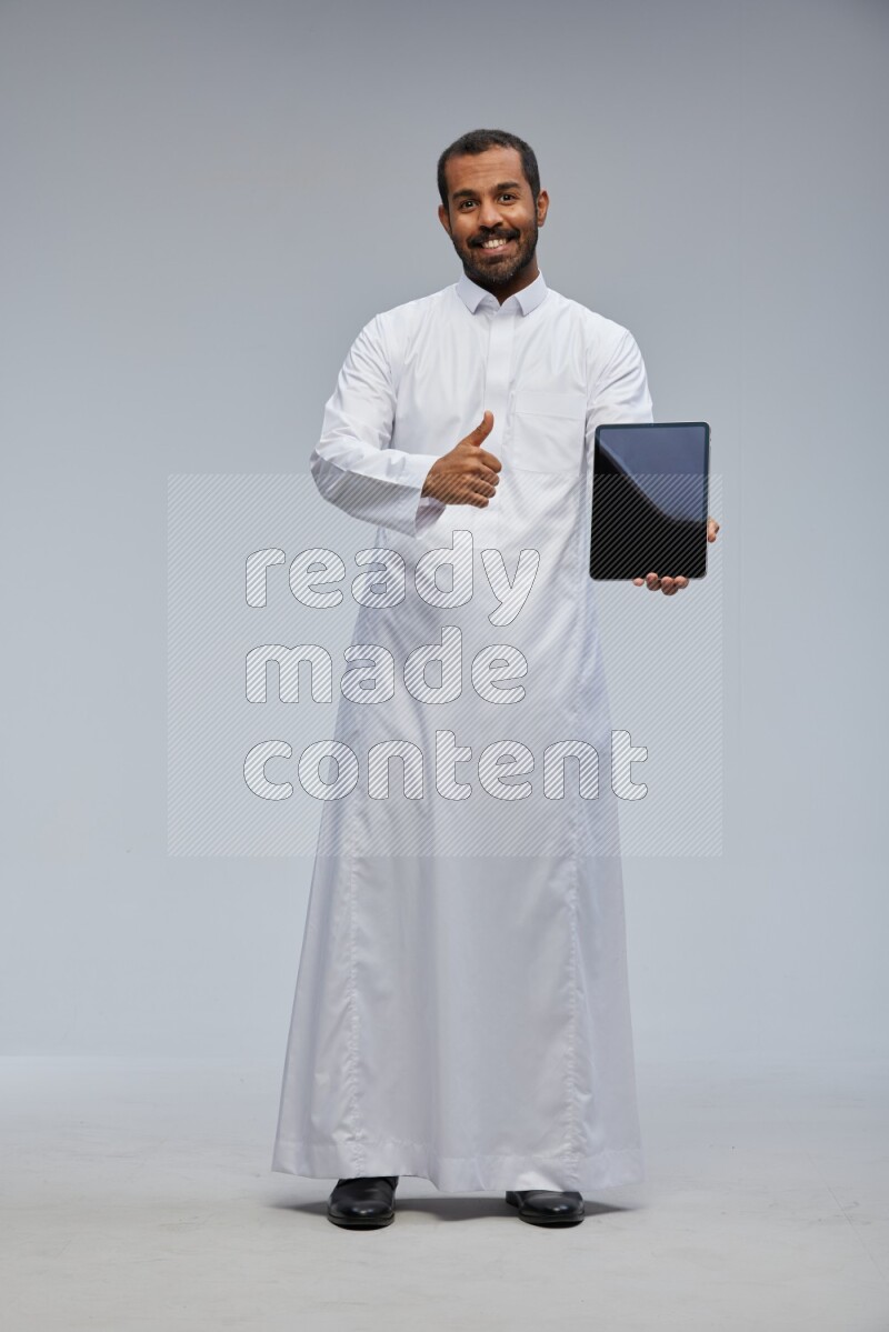 Saudi man wearing Thob standing showing tablet to camera on Gray background