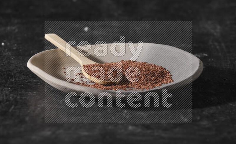 A multicolored pottery plate full of garden cress seeds and wooden spoon full of seeds on a textured black flooring