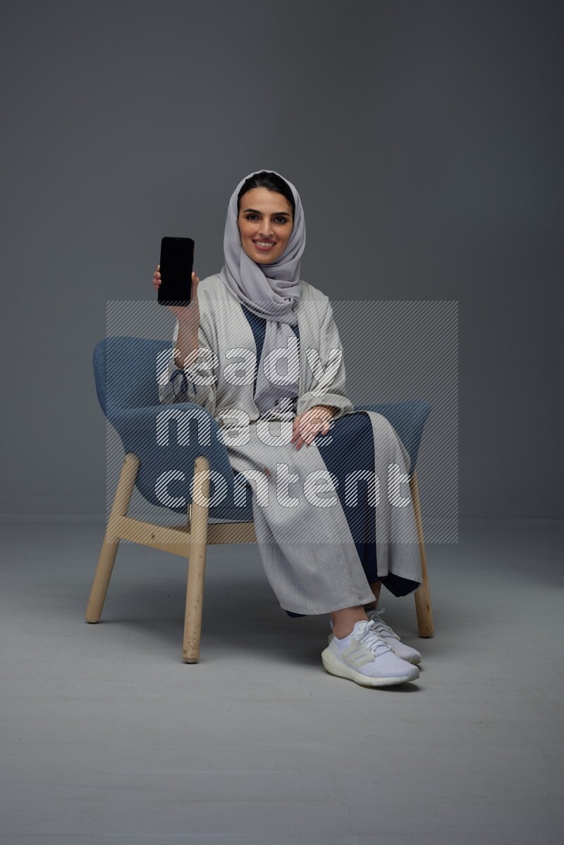 A Saudi woman wearing a light gray Abaya and head scarf sitting on a dark grey chair and showing the phone's screen eye level on a grey background