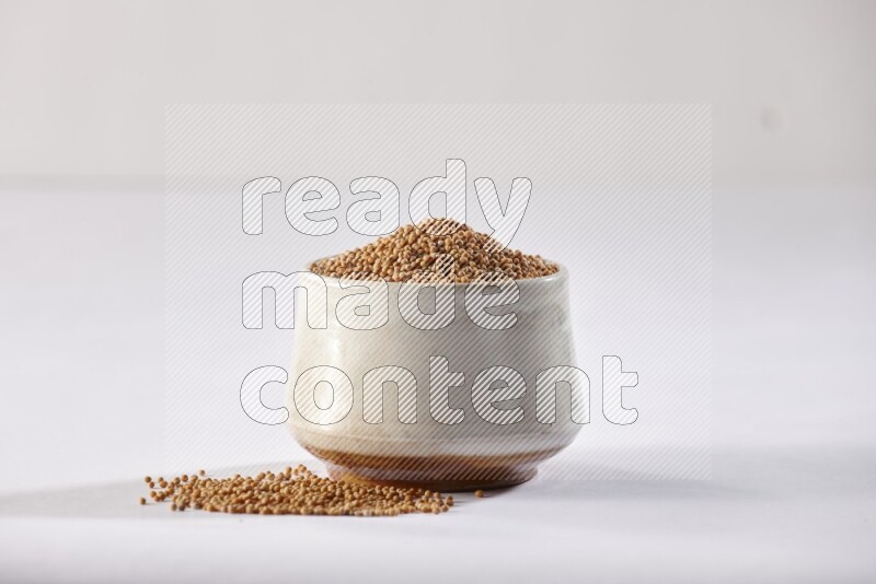 A beige pottery bowl full of mustard seeds and more seeds spread on a white flooring