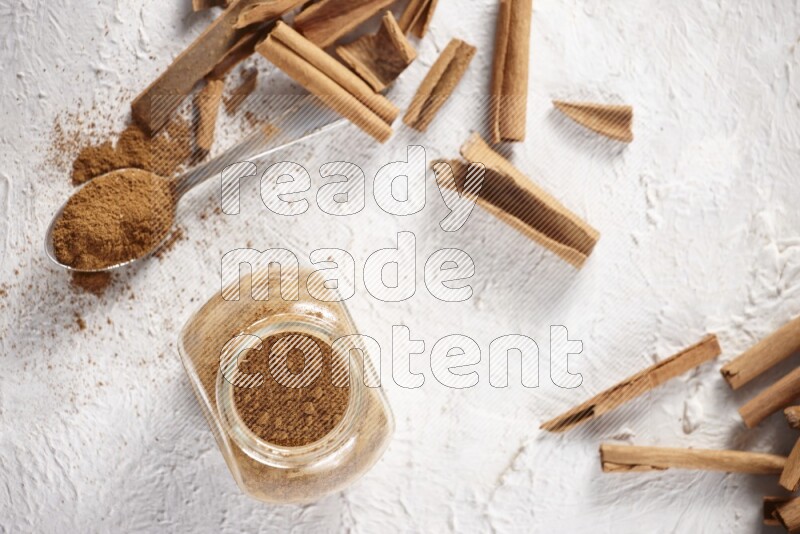 Herbal glass jar full cinnamon powder and a metal spoon surrounded by cinnamon sticks on a white background