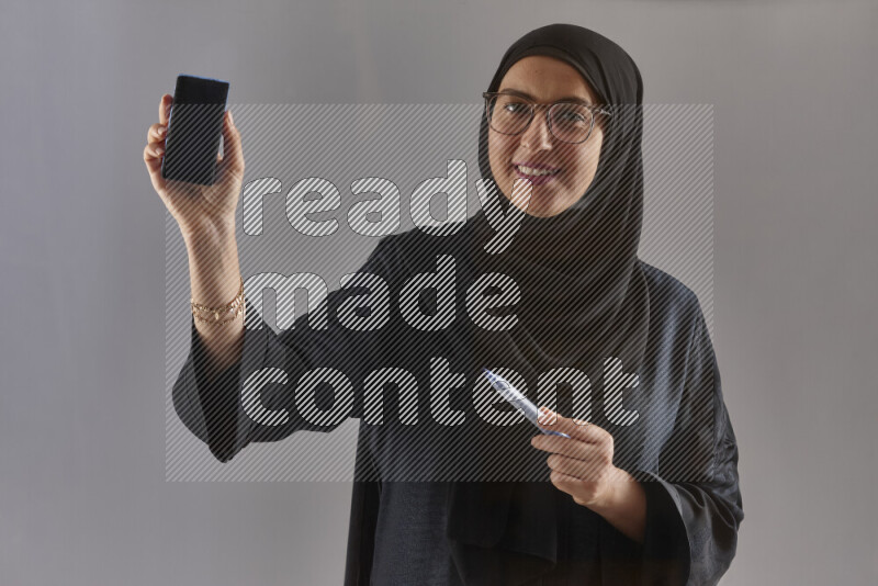 Woman in black abaya, hijab and glasses holding a marker pen to write on a transparent board with different reactions and expressions on grey background