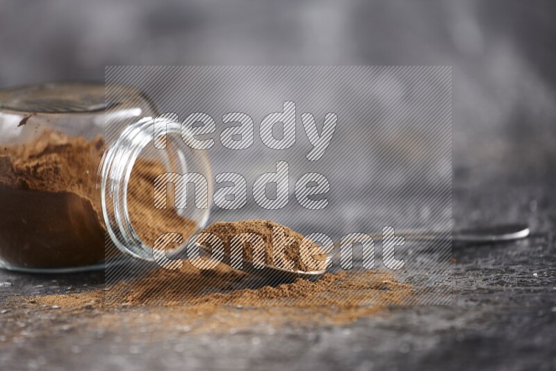 Herbal glass jar full of cinnamon powder flipped and a metal spoon on textured black background