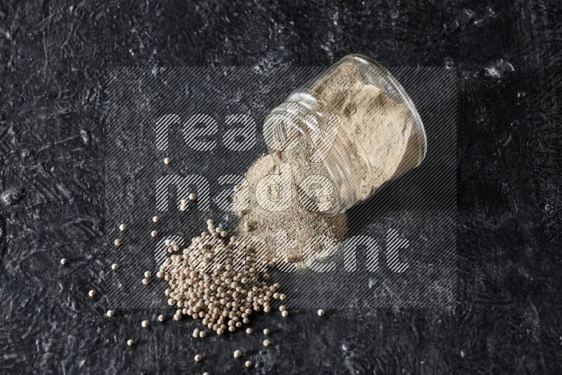 A flipped glass jar full of white pepper powder with spilled powder and pepper beads on textured black flooring