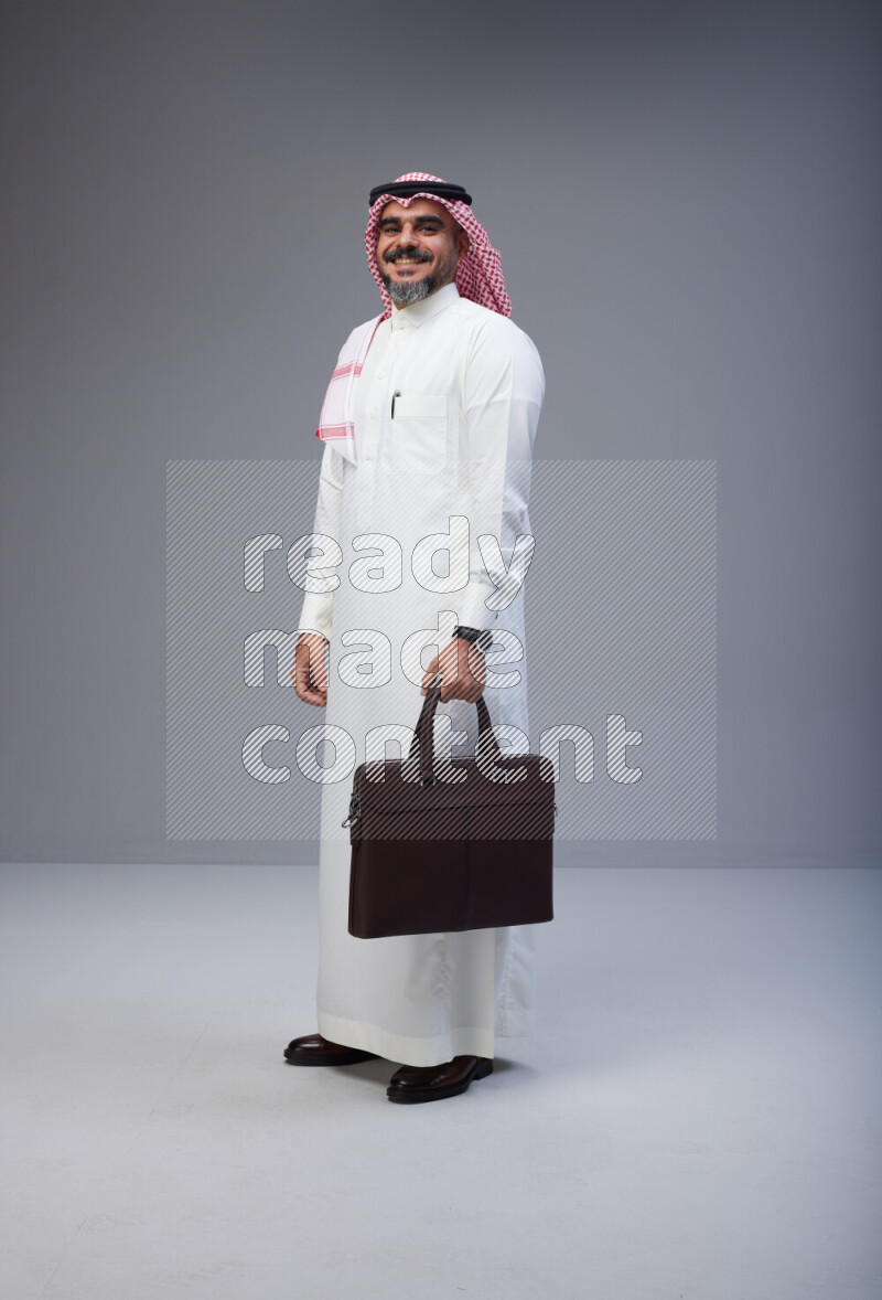 Saudi man Wearing Thob and red Shomag standing holding bag on Gray background