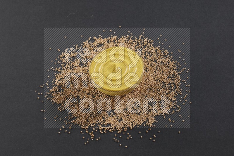 A glass bowl full of mustard paste with mustard seeds underneath on black flooring