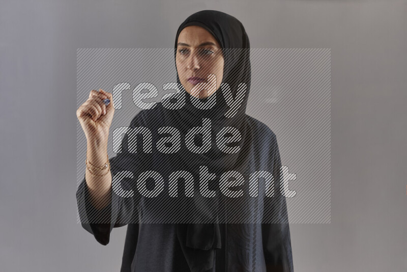 Woman in black abaya and hijab holding a marker pen to write on a transparent board with different reactions and expressions on grey background