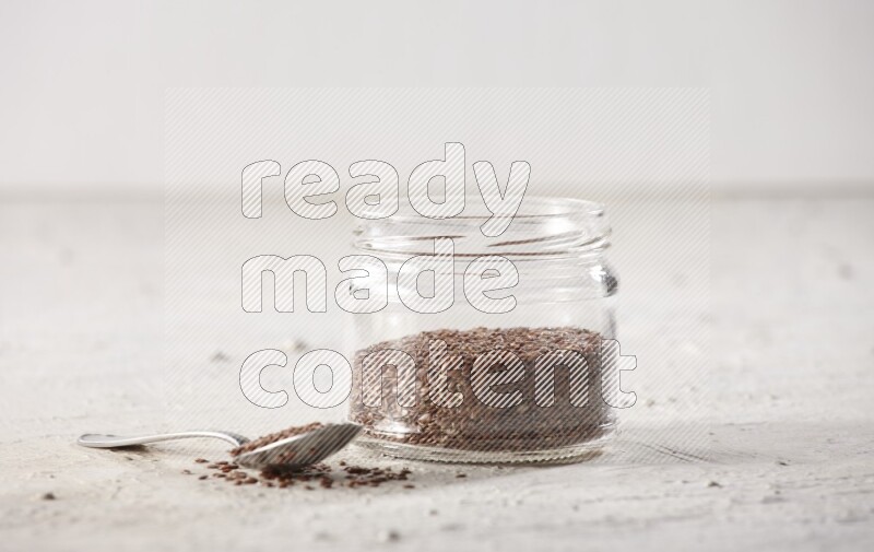 A glass jar full of flax seeds with a metal spoon full of the seeds on a textured white flooring