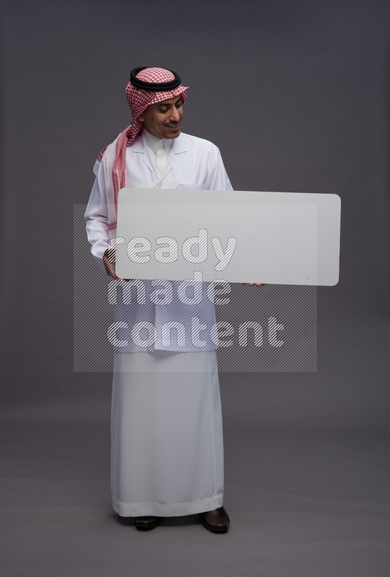 Saudi man wearing thob with lab coat and shomag with pocket employee badge standing holding board on gray background