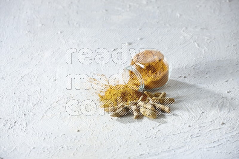 A flipped glass spice jar full of turmeric powder and powder spilled out of it with dried whole fingers on textured white flooring