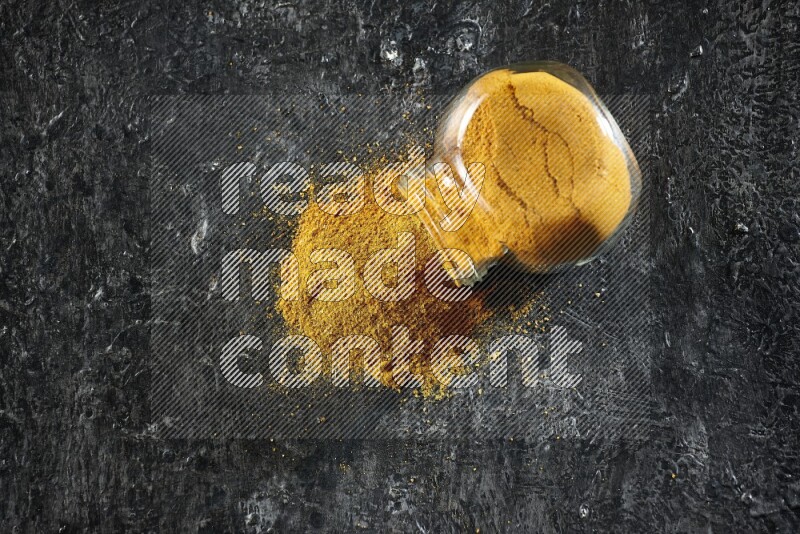 A flipped glass spice jar full of turmeric powder and powder spilled out of it on textured black flooring