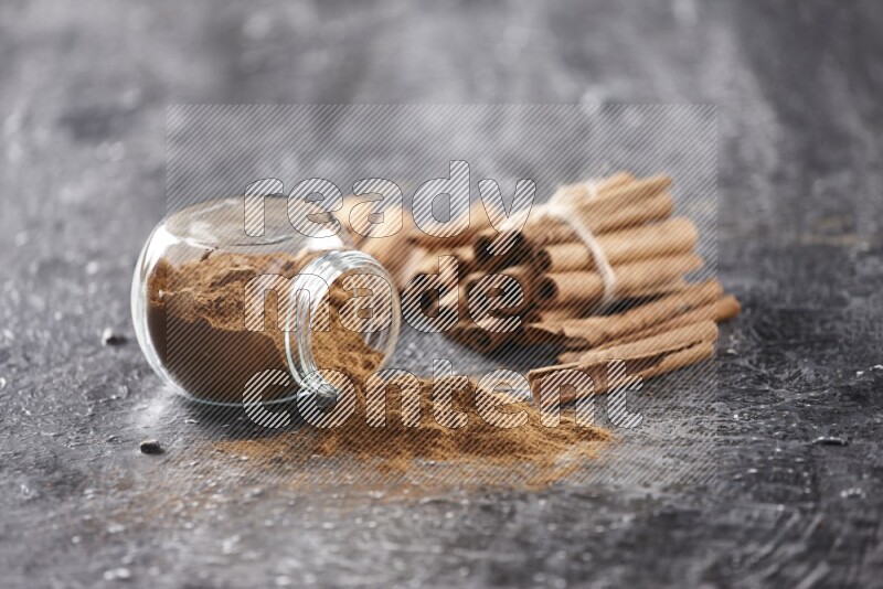 Herbal glass jar full of cinnamon powder flipped with cinnamon sticks stacked and bounded on a textured black background