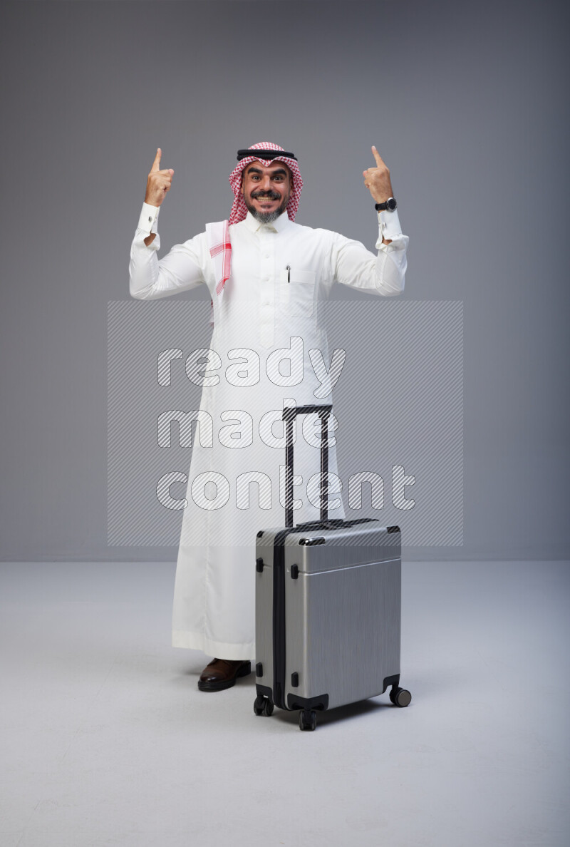 Saudi man wearing Thob and red Shomag standing holding Travel bag on Gray background
