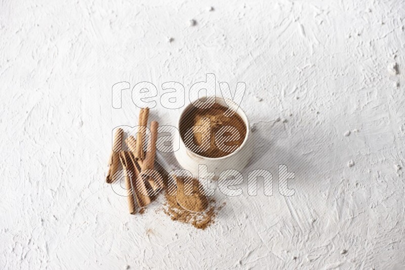 Ceramic beige bowl full of cinnamon powder and a metal spoon with cinnamon sticks next of it on a textured white background