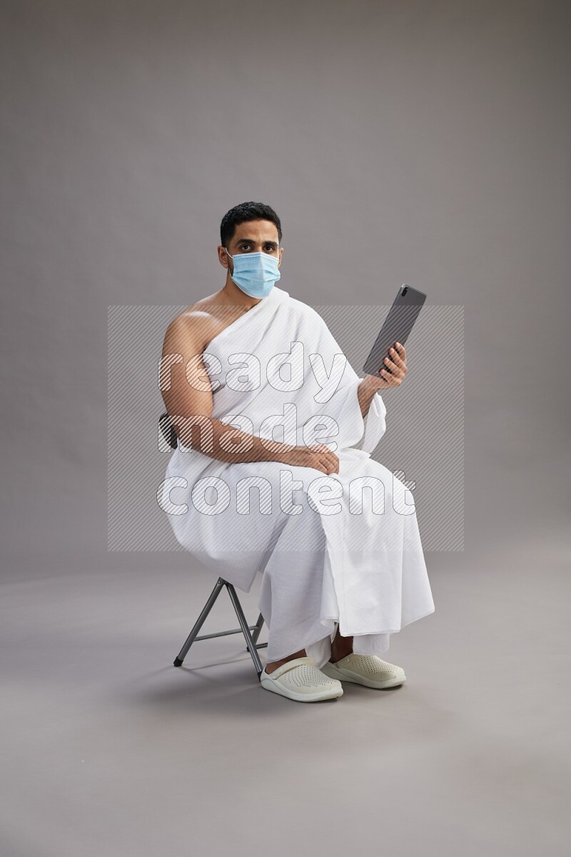 A man wearing Ehram with face mask sitting on chair holding a tablet on gray background