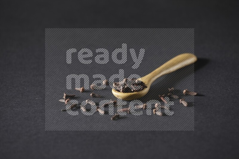 A wooden spoon full of cloves and some spreaded out of it on a black flooring
