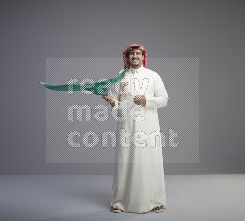 A Saudi man standing wearing thob and red shomag raising big Saudi flag on gray background