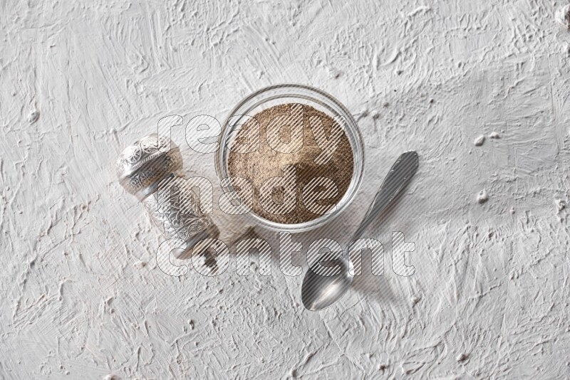 A glass bowl full of black pepper powder and a metal spoon and a turkish metal grinder on textured white flooring