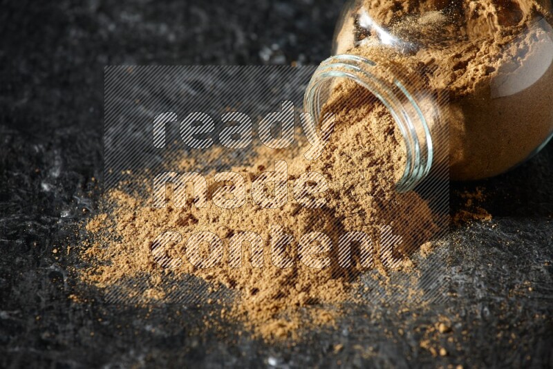 A flipped glass spice jar full of allspice powder and powder spilled out of it on a textured black flooring