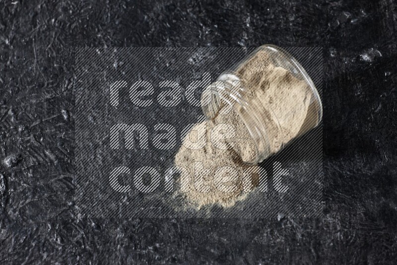 A flipped glass jar full of white pepper powder with spilled powder on textured black flooring