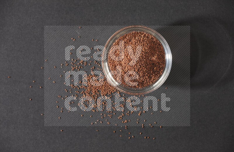 A glass bowl full of garden cress seeds surrounded by seeds on a black flooring